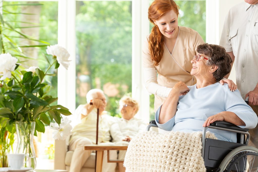 Tender caregiver saying goodbye to an elderly pensioner in a wheelchair in a day care facility A companion pushing the wheelchair Other elderly people in the blurred background